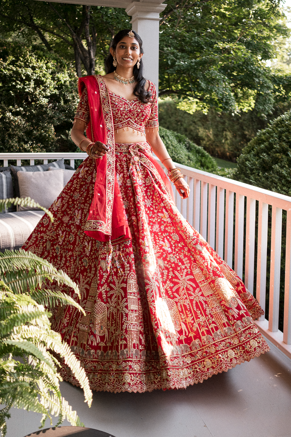 A woman in a red and gold traditional Indian bridal outfit, standing on a porch with greenery in the background, smiling at the camera.