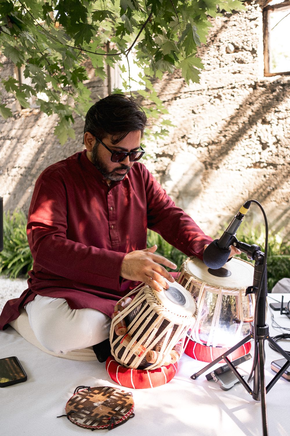 A man wearing sunglasses and a maroon kurta playing two drums with a microphone placed nearby in an outdoor setting with leafy trees and a rustic stone wall in the background.