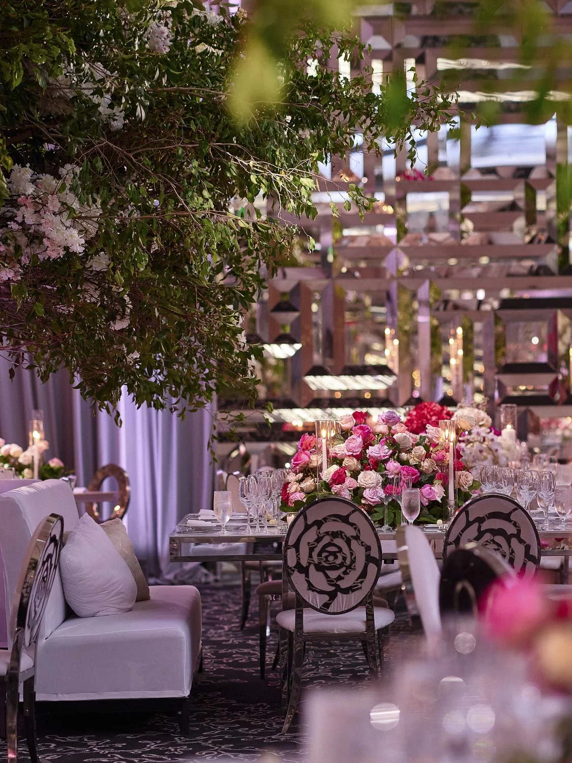 Elegant event space with a large floral centerpiece of pink and white roses, surrounded by glassware and candles, and decorated with lush greenery and pink lighting.