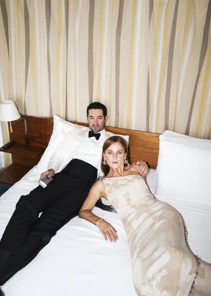 A man in a black tuxedo with a bowtie and a woman in an elegant beige dress lying on a hotel bed with striped wallpaper background.