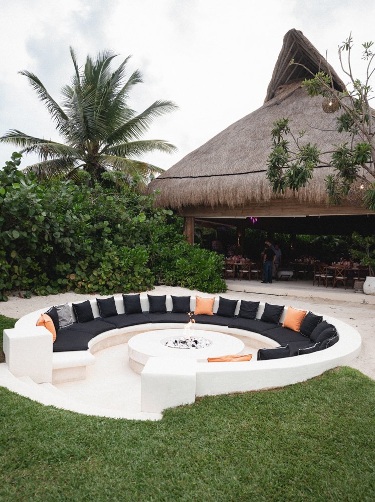Outdoor seating area with a circular white concrete bench with black and peach cushions around a fire pit, with tropical vegetation and a thatched-roof open-air restaurant in the background.