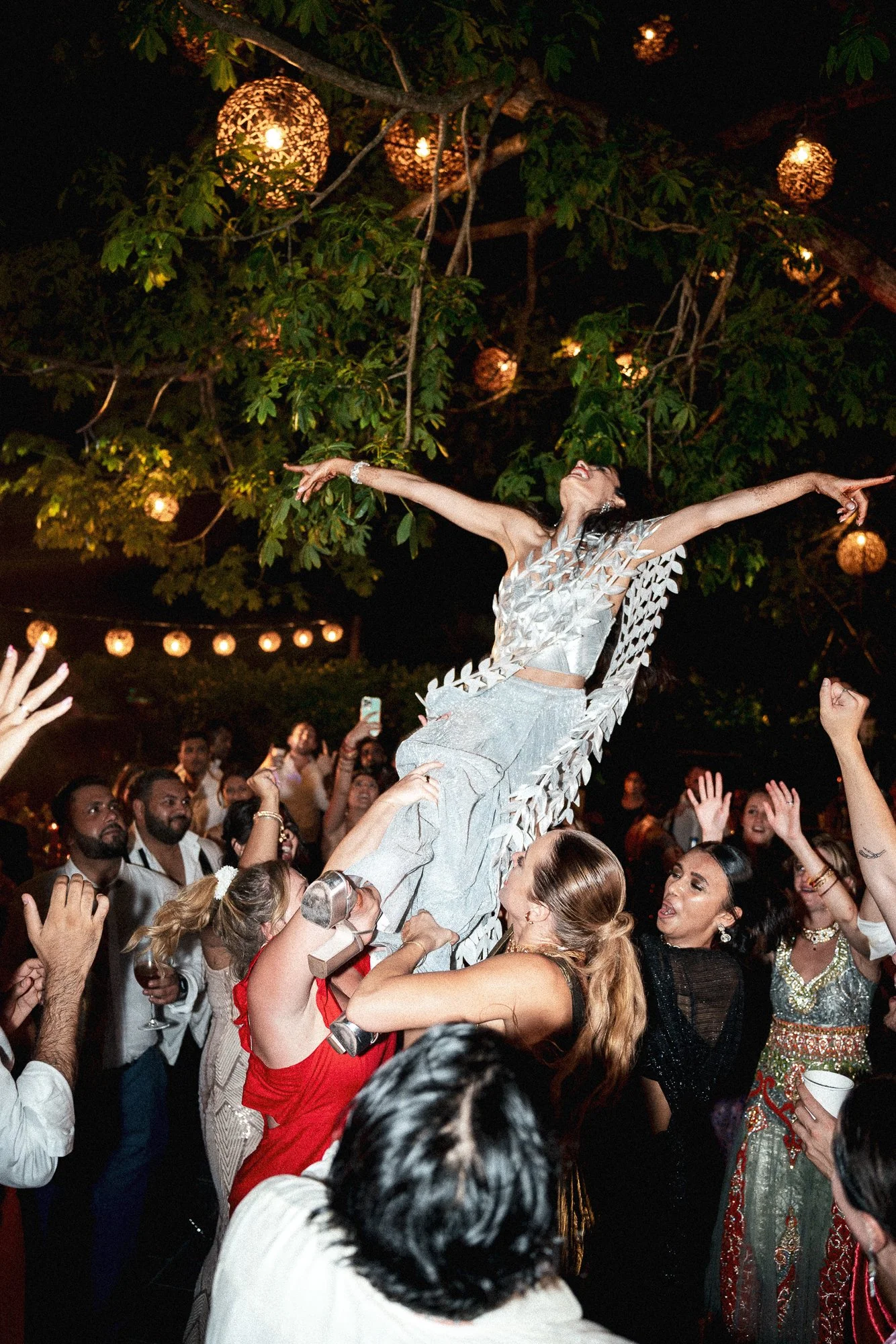 A woman being lifted above a crowd at a nighttime celebration, reaching with arms outstretched, under decorative hanging lights and surrounded by smiling people.
