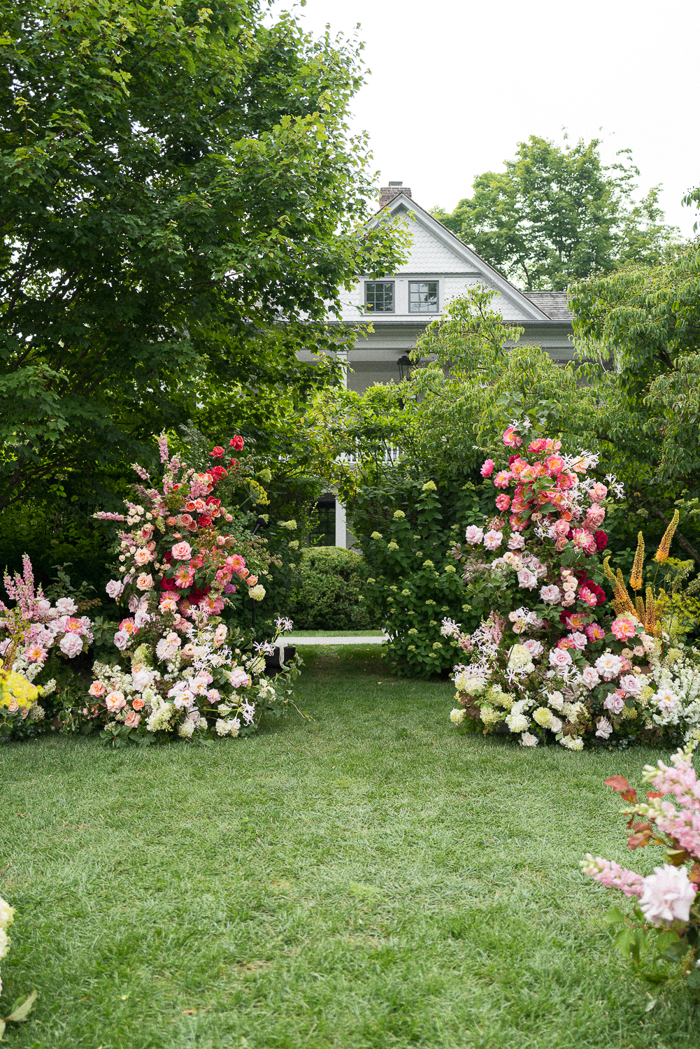 A garden with lush green trees and vibrant pink and white flower arrangements on either side of a pathway, with a house partially visible in the background.