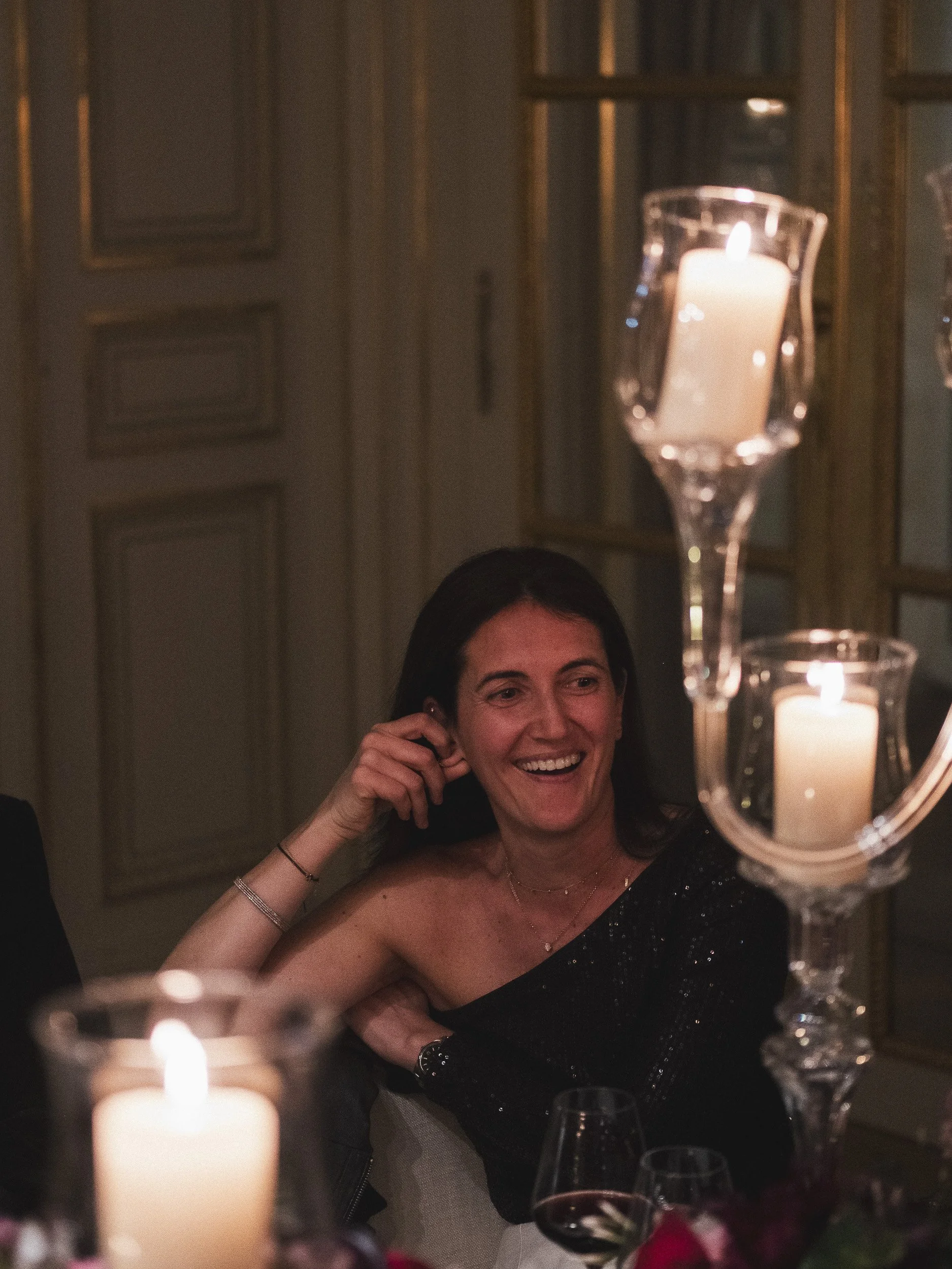 A woman smiling at a dinner party, with candles and wine glasses on the table.