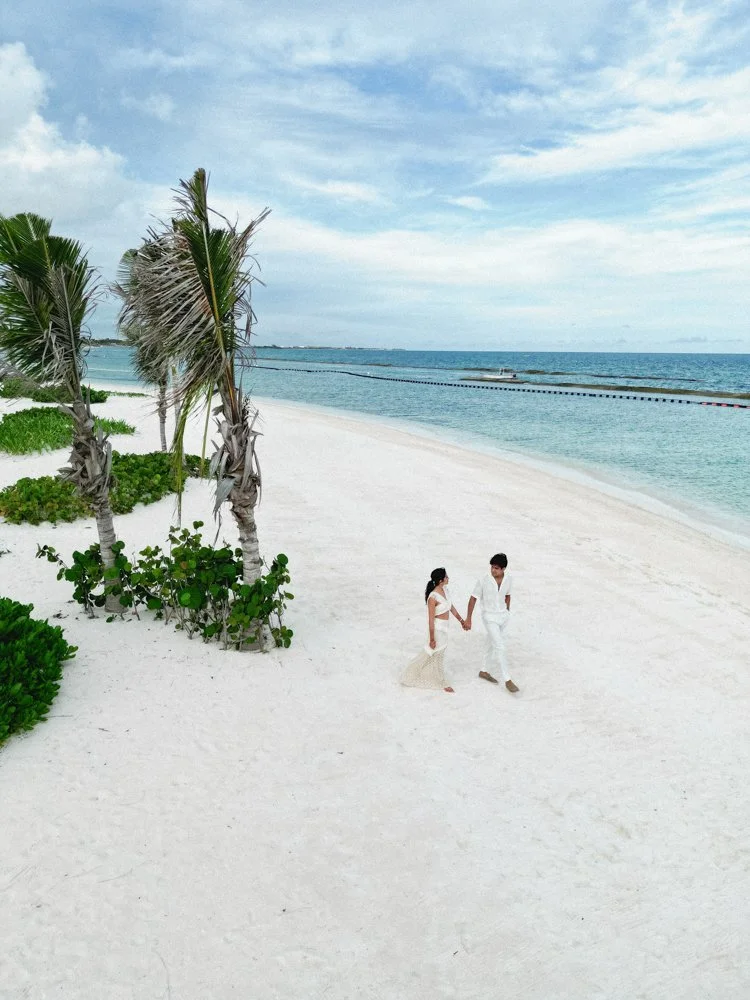 A couple walking hand in hand on a sandy beach with palm trees and ocean in the background.