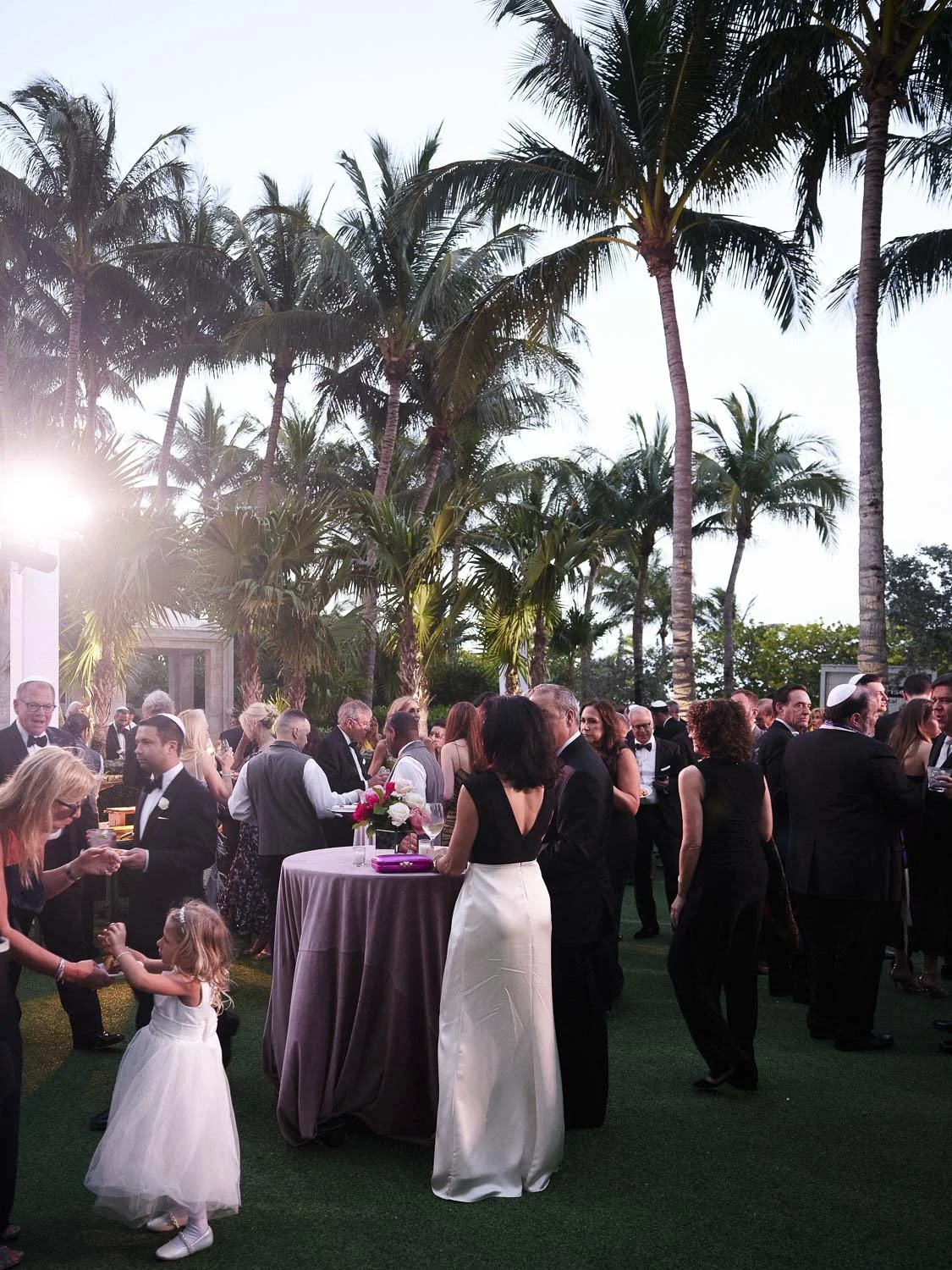 Guests at an outdoor wedding reception under palm trees, mingling and dancing during sunset.