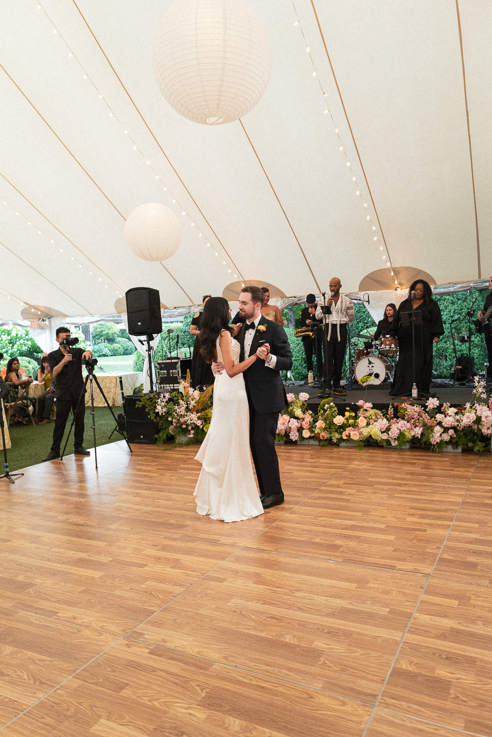 Couple dancing at their wedding reception under a large tent with string lights, a band playing in the background, and guests watching.
