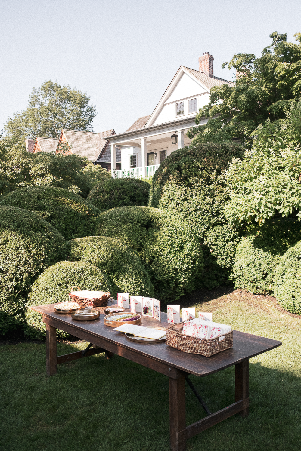Table set up outdoors with various items, surrounded by large bushes, in front of a white house with a porch and green trees.