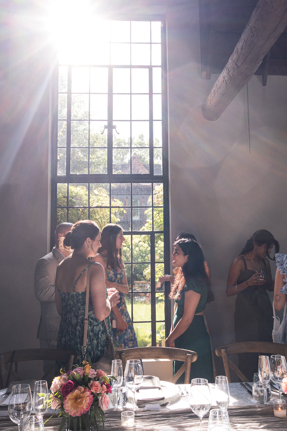 Group of women and one man talking and mingling inside a sunny room with large window, decorated table with flowers and glasses in the foreground.