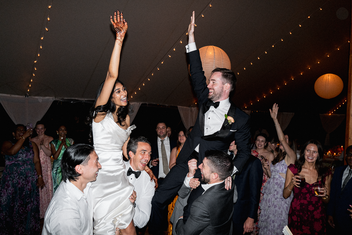 Groom and bride lifting their hands in celebration during a wedding reception, surrounded by guests under string lights and paper lanterns.