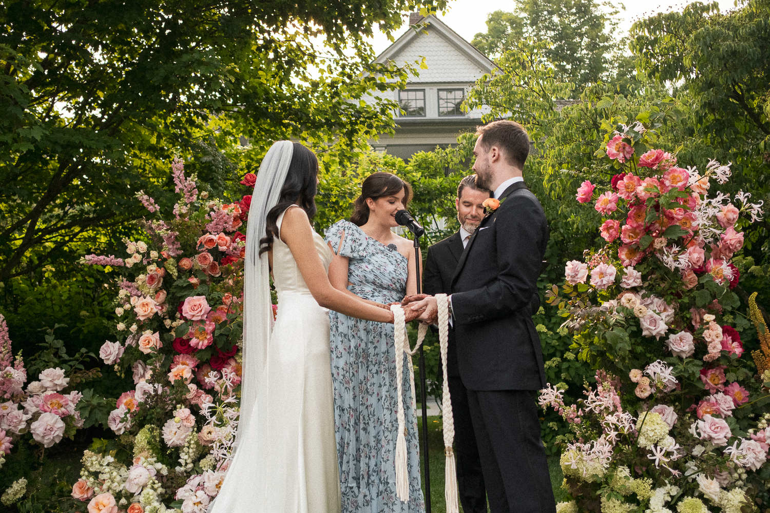 A couple getting married outdoors, holding hands, with a officiant and wedding officiant standing nearby, surrounded by pink and white flowers and greenery.