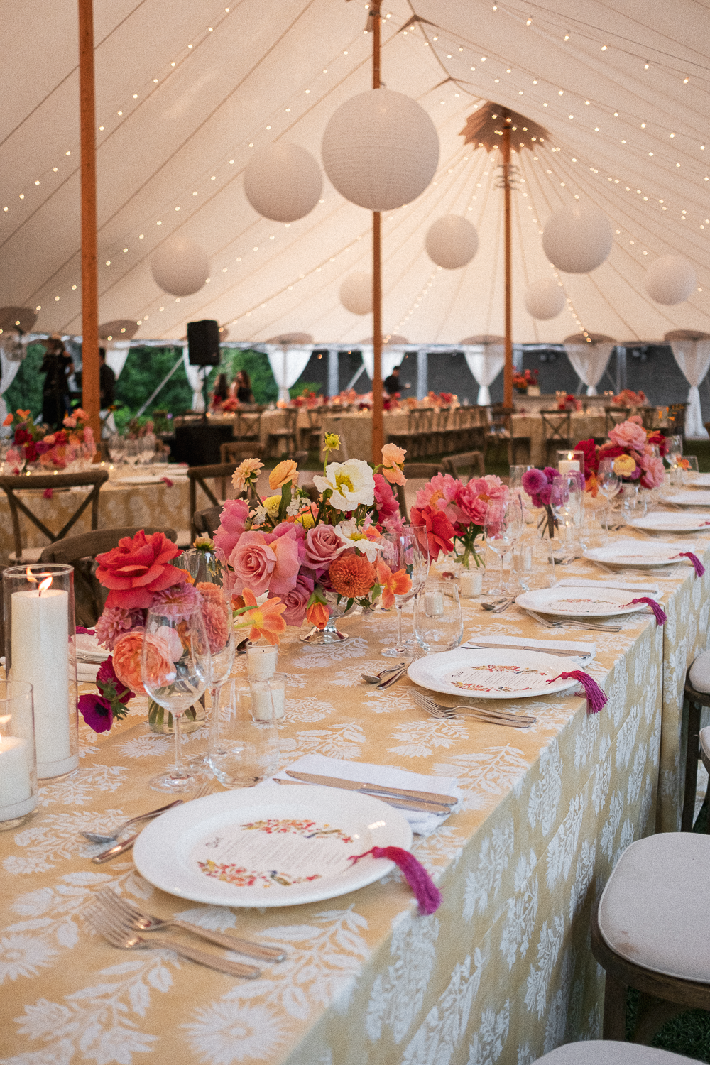 A beautifully decorated outdoor event tent featuring a long table with pink and peach floral arrangements, candles, and elegant table settings, illuminated by string lights hanging from the ceiling.
