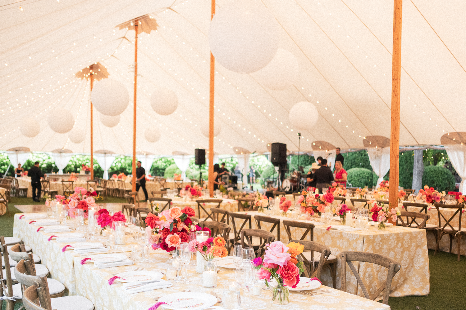 A decorated wedding reception tent with long tables covered in yellow floral tablecloths, pink and peach floral centerpieces, surrounded by wooden chairs, and a band setting up in the background.