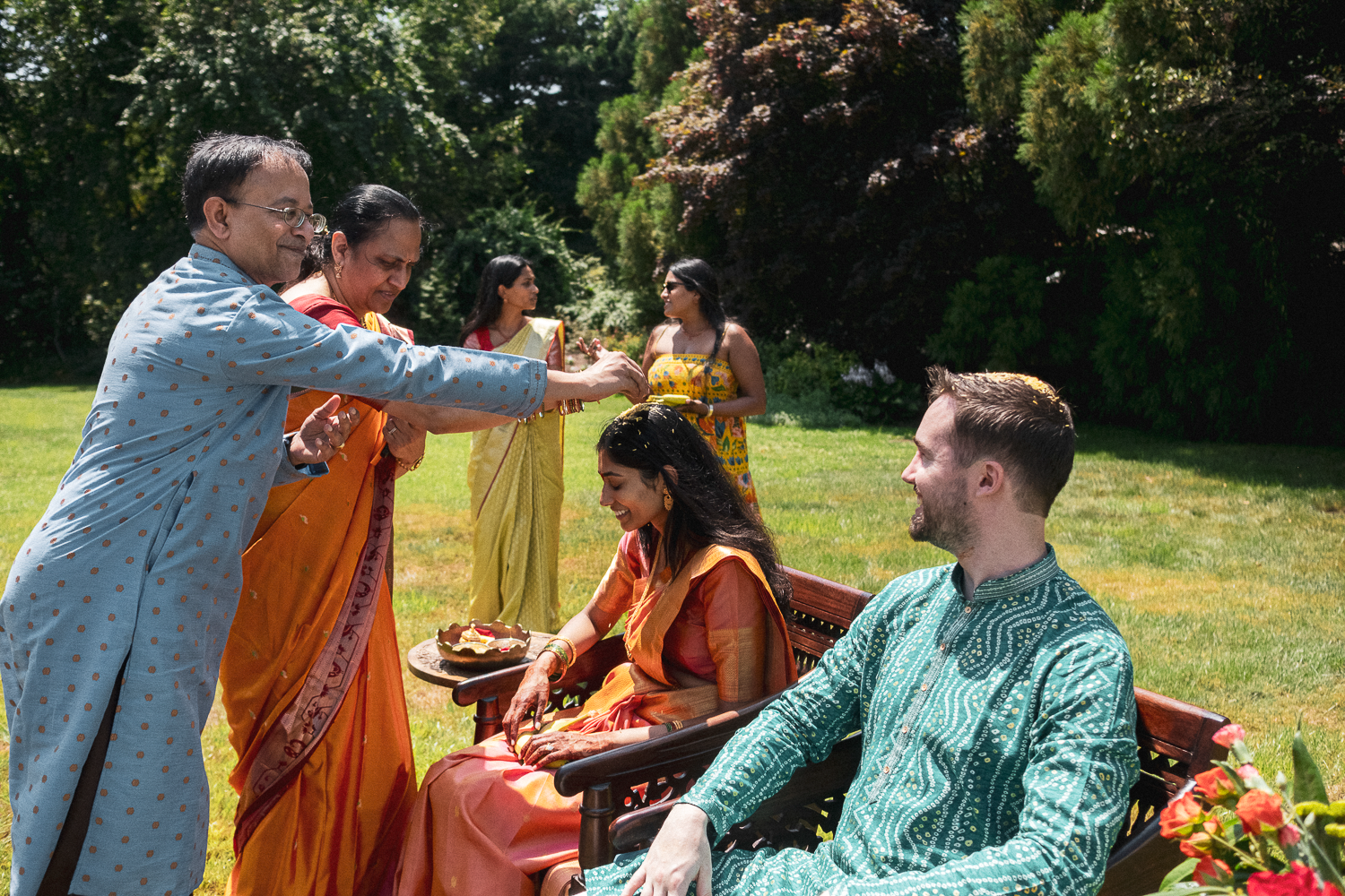 Indian woman performing a traditional ceremony on a smiling Caucasian man sitting on a bench outdoors during daytime, with two women in traditional attire standing behind them.