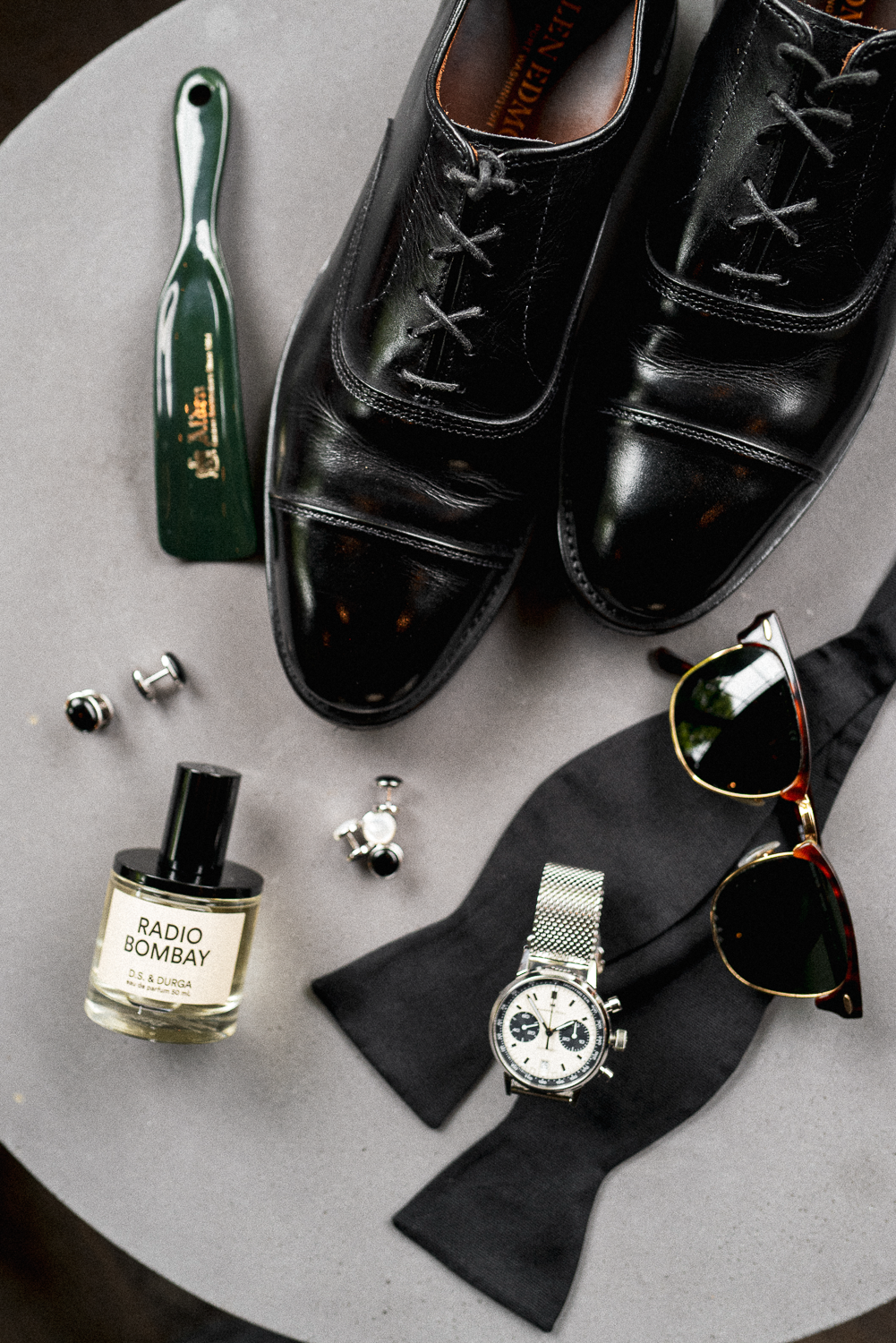 Black leather dress shoes, sunglasses with gold frames, a watch with a metal strap, cufflinks, an aftershave bottle labeled 'RADIO BOMBAY,' a black bow tie, and a green shoehorn on a gray round surface.