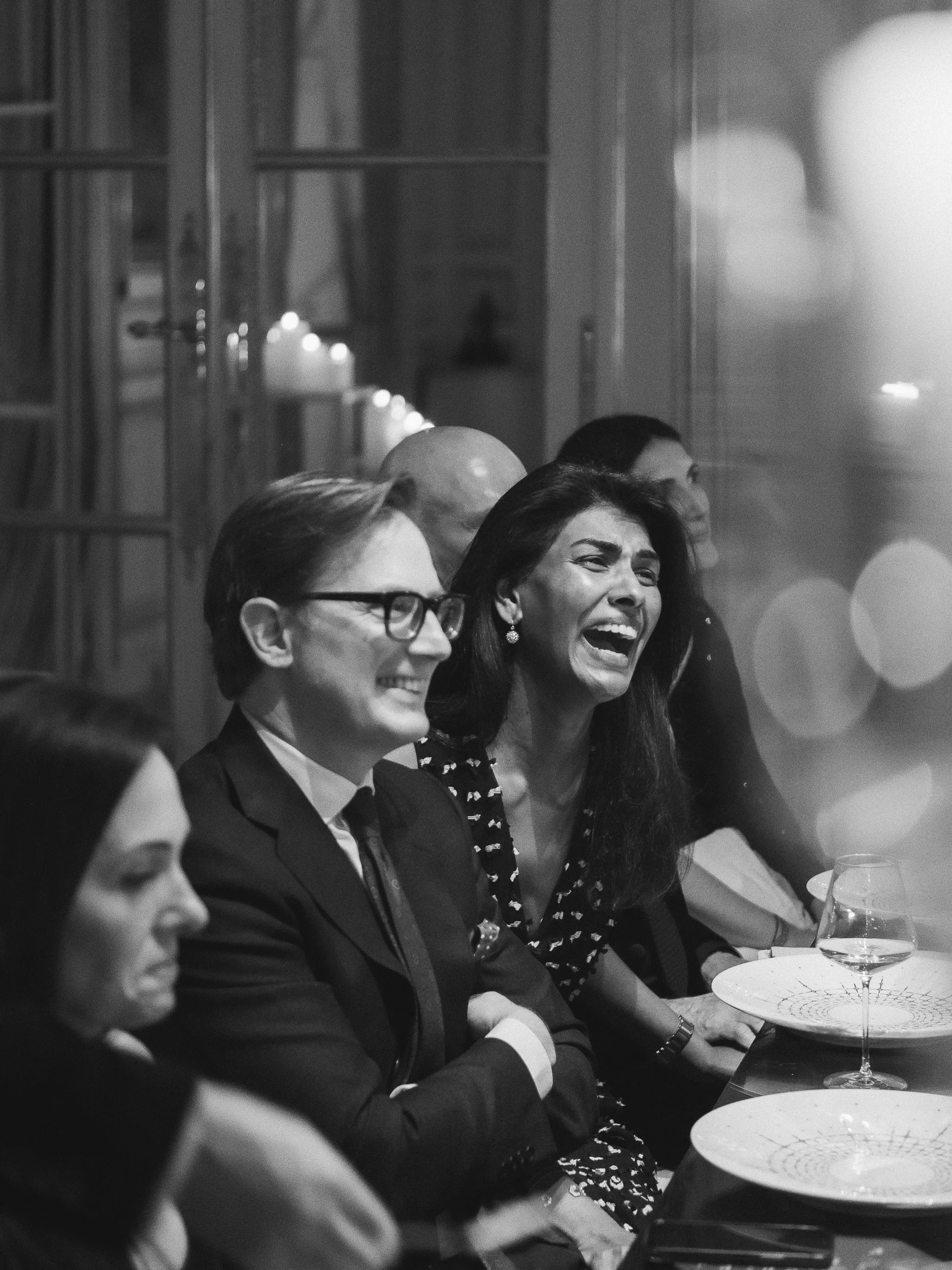People sitting at a table, laughing and smiling, during a formal event, with candles and glassware on the table, in a room with wooden shelves in the background.
