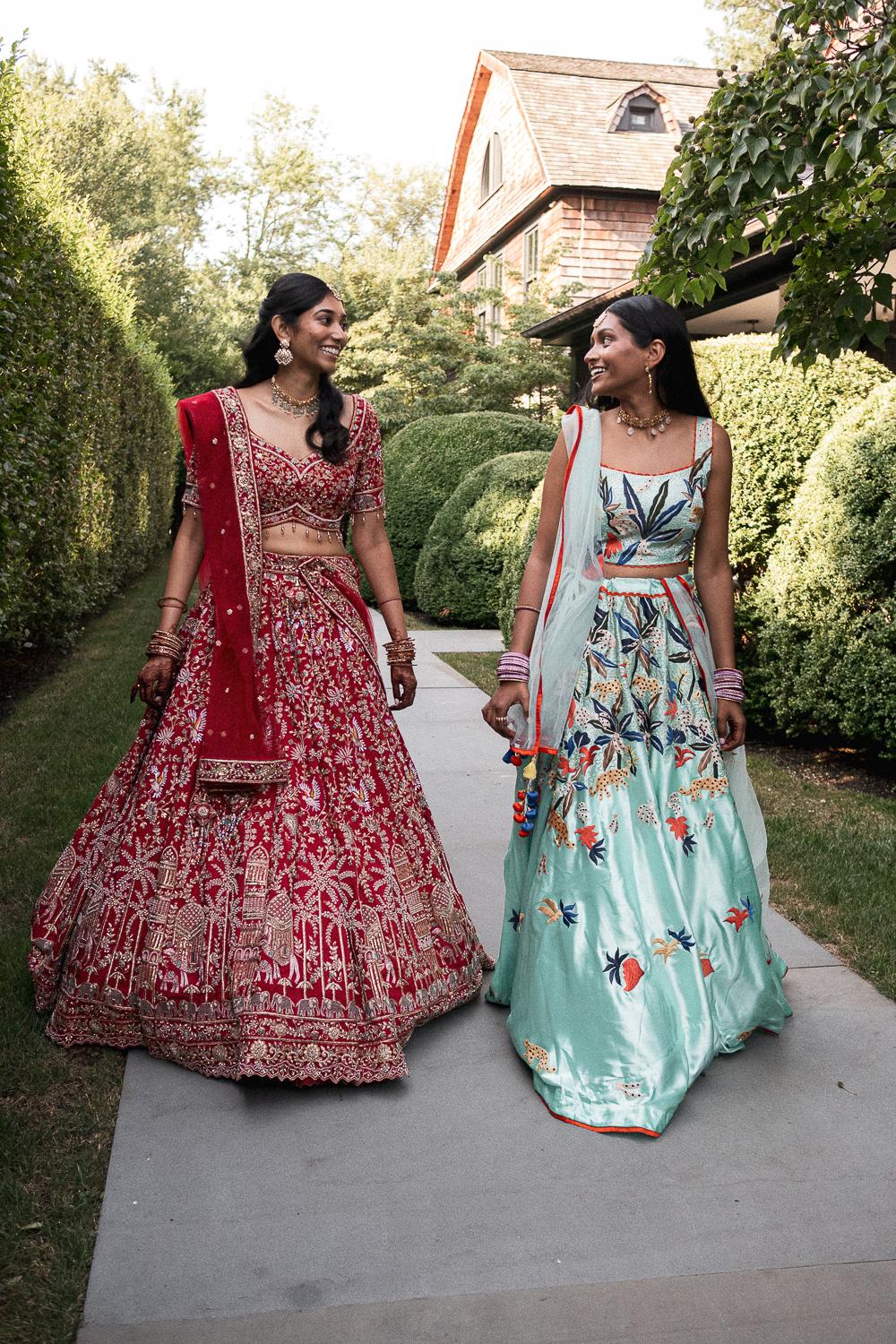 Two women wearing traditional Indian dresses walking on a sidewalk outdoors, surrounded by green bushes and trees, with a house in the background.