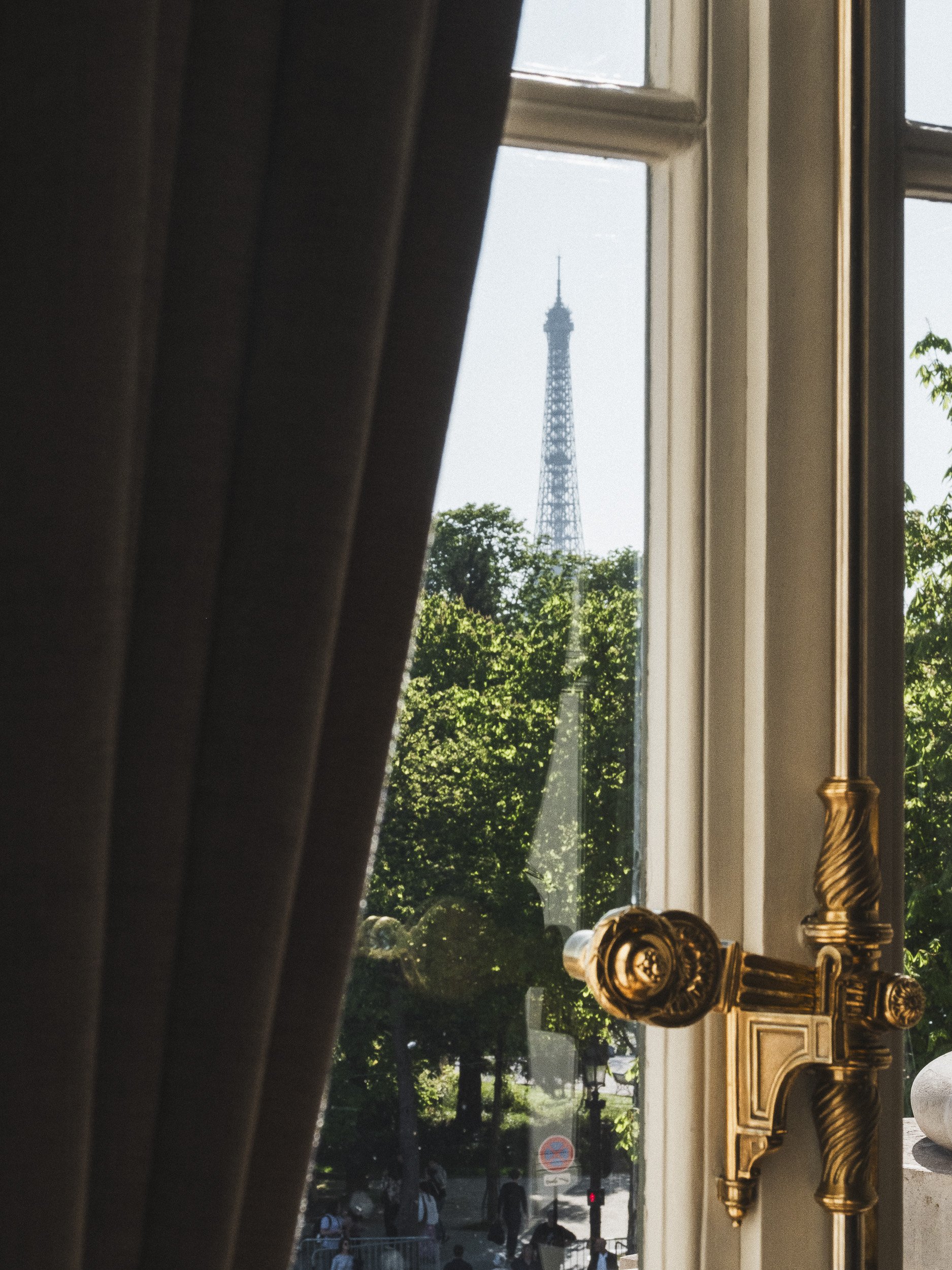 View of Eiffel Tower through a window with partially drawn curtains, gold-colored curtain tiebacks, and a view of a city street with trees and pedestrians outside.