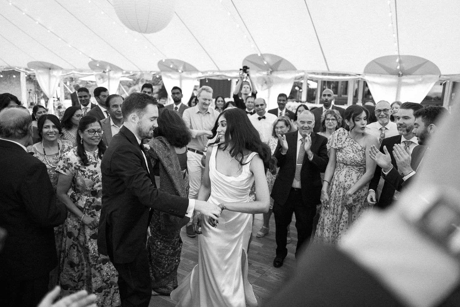 A bride and groom dancing at their wedding reception with guests watching and clapping in the background inside a decorated tent.