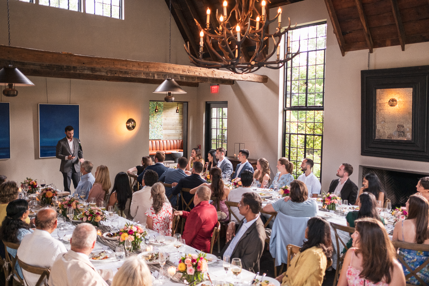 A large group of people seated at a long dining table during an event in a spacious room with large windows, decorative lighting, and wooden accents, listening to a man in a suit speaking at the front.