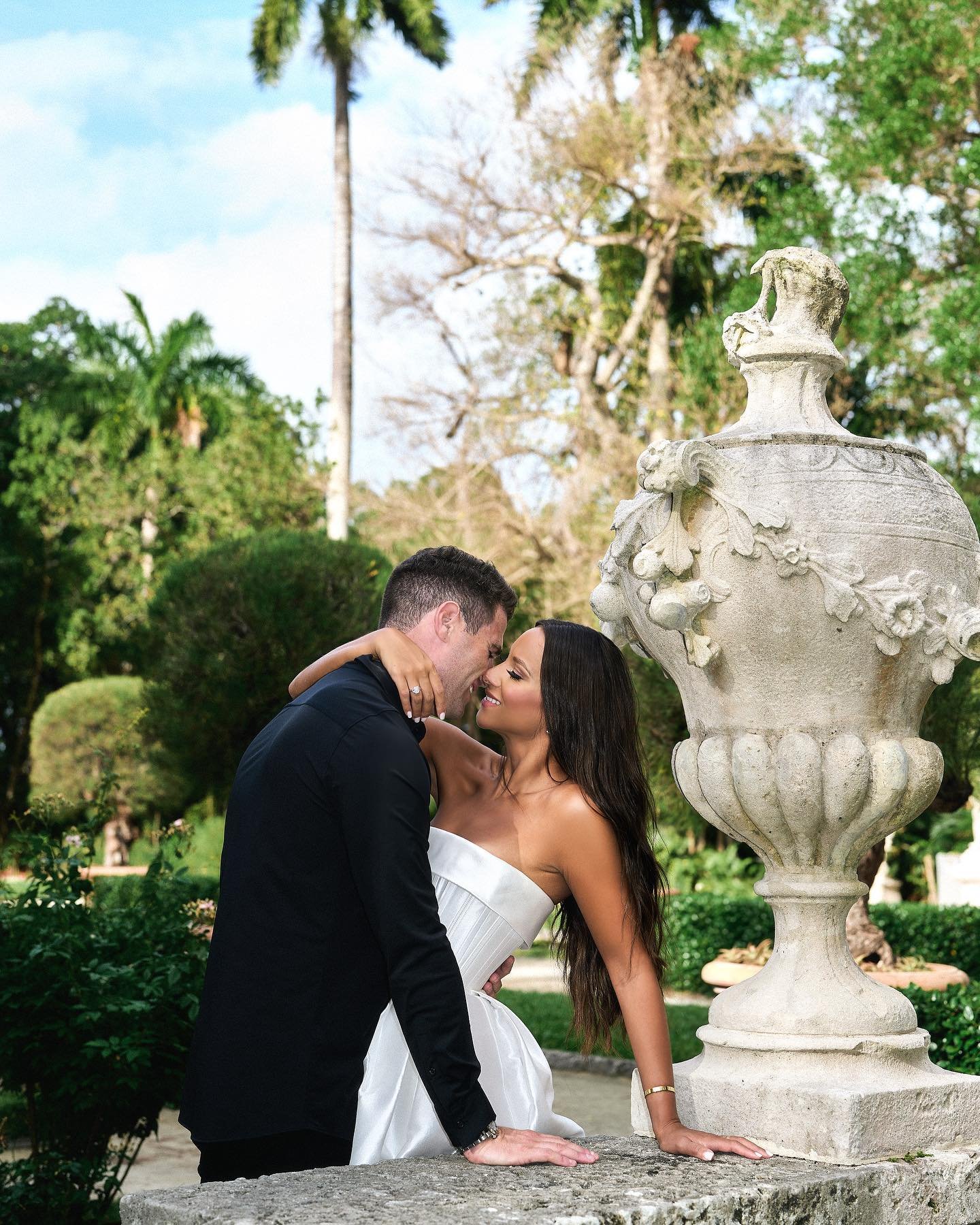 A couple dressed in formal attire, with the man in a black suit and the woman in a white strapless gown, are sharing an intimate moment outdoors near a decorative stone fountain, surrounded by greenery and trees.