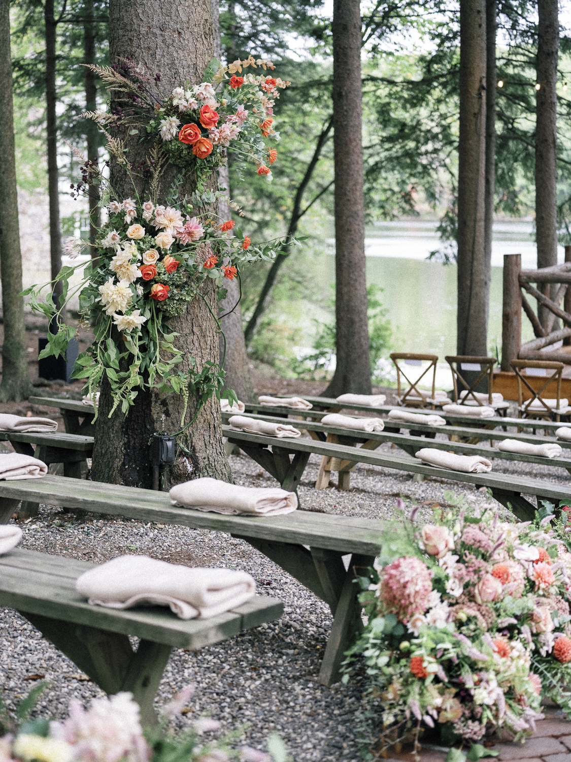 Outdoor wedding ceremony setup with benches and floral arrangements, decorated with pink, white, and orange flowers, in a wooded area beside a body of water.