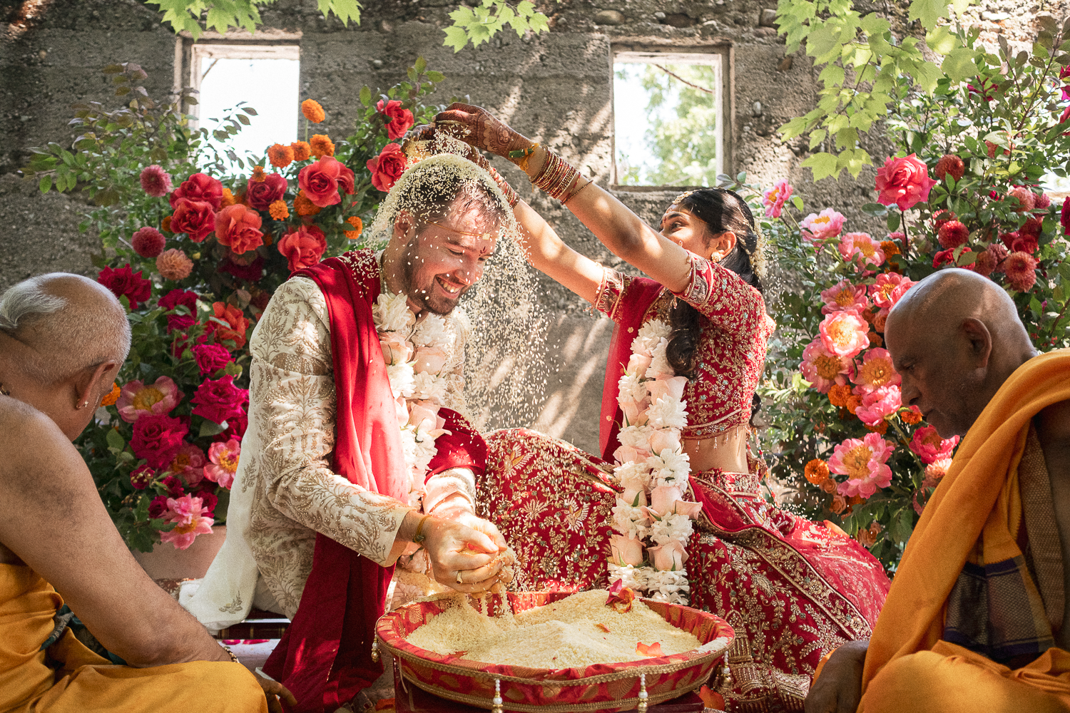 Indian wedding ceremony with a bride and groom performing a traditional ritual, surrounded by flower arrangements, with priests and floral decorations.