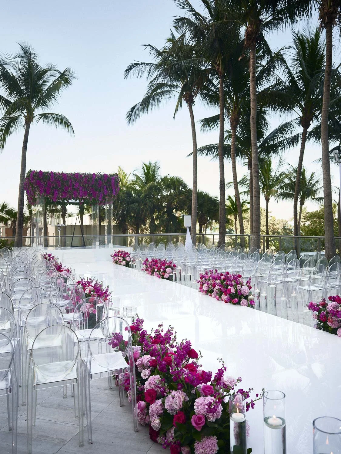Outdoor wedding venue decorated with pink and purple flowers, transparent chairs, and palm trees with a clear sky.