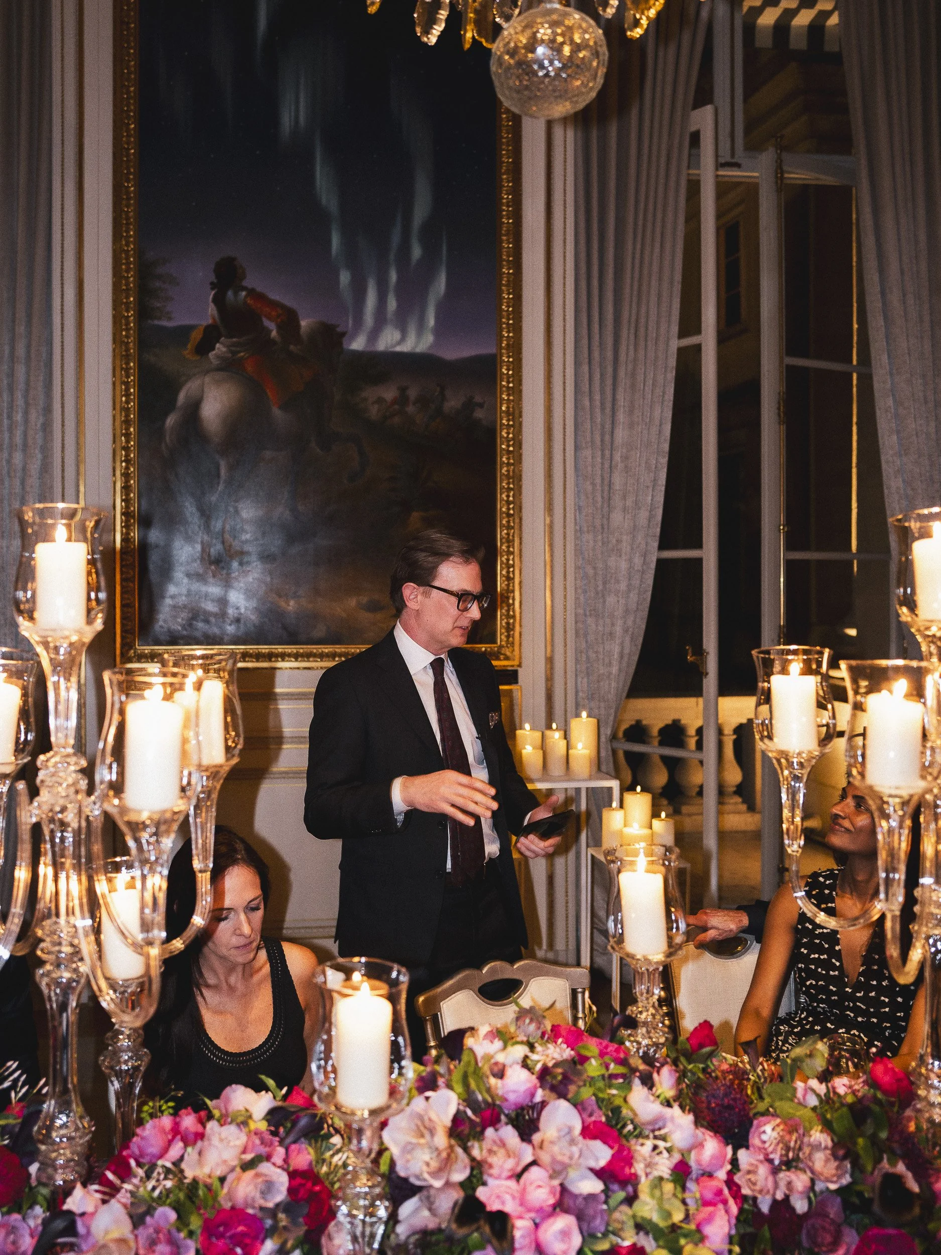 A formal dinner party in an elegant room with tall windows, curtains, and a chandelier. The table is decorated with numerous candles and a large arrangement of pink and purple flowers. Several people are seated and one man is standing, talking to the