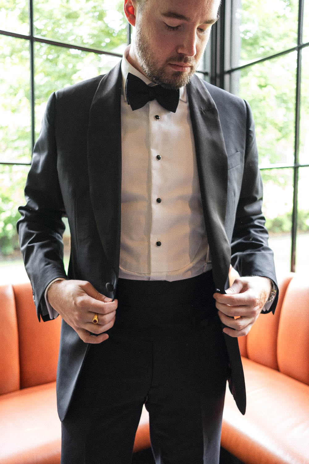 A man wearing a tuxedo with a white dress shirt, black bow tie, and black jacket, standing indoors near a large window with greenery outside.