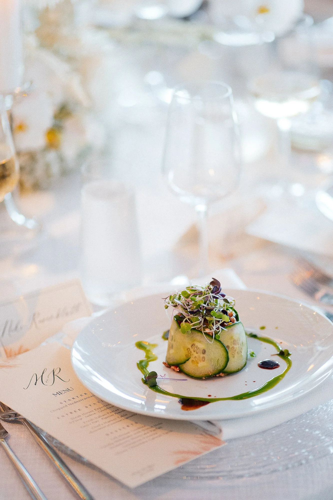 A gourmet appetizer dish on a white plate, garnished with microgreens and cucumber slices, drizzled with green and dark sauce, placed on a table with a menu and a clear glass of water in the background, at a fancy dining setting.