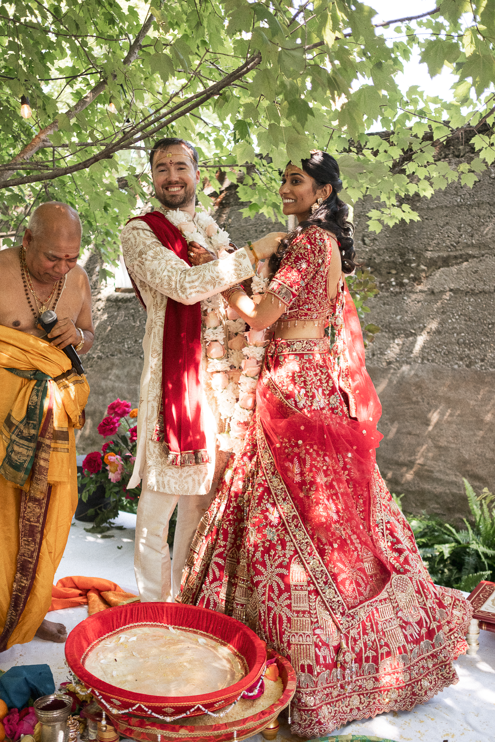 Indian wedding ceremony with a bride and groom exchanging garlands under a leafy tree, officiant standing nearby, and flower decorations.