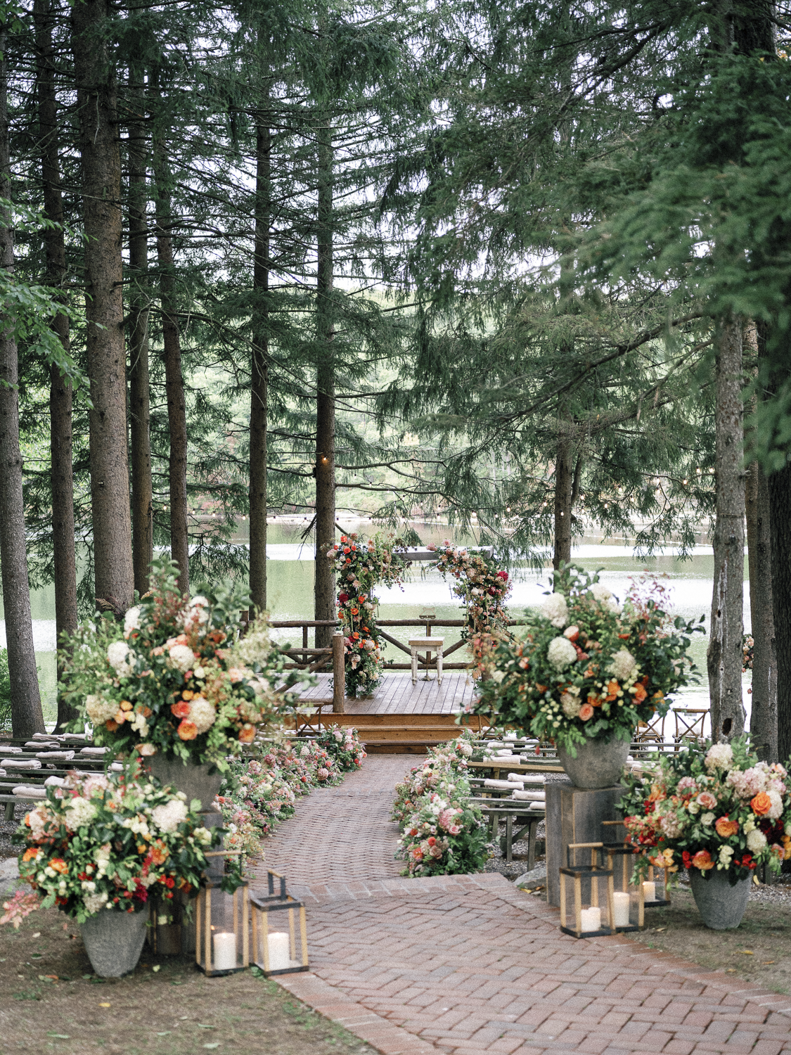 Outdoor wedding setup with a wooden platform decorated with an arch of flowers, surrounded by large floral arrangements, lanterns, and candles, in a forested area by a lake.
