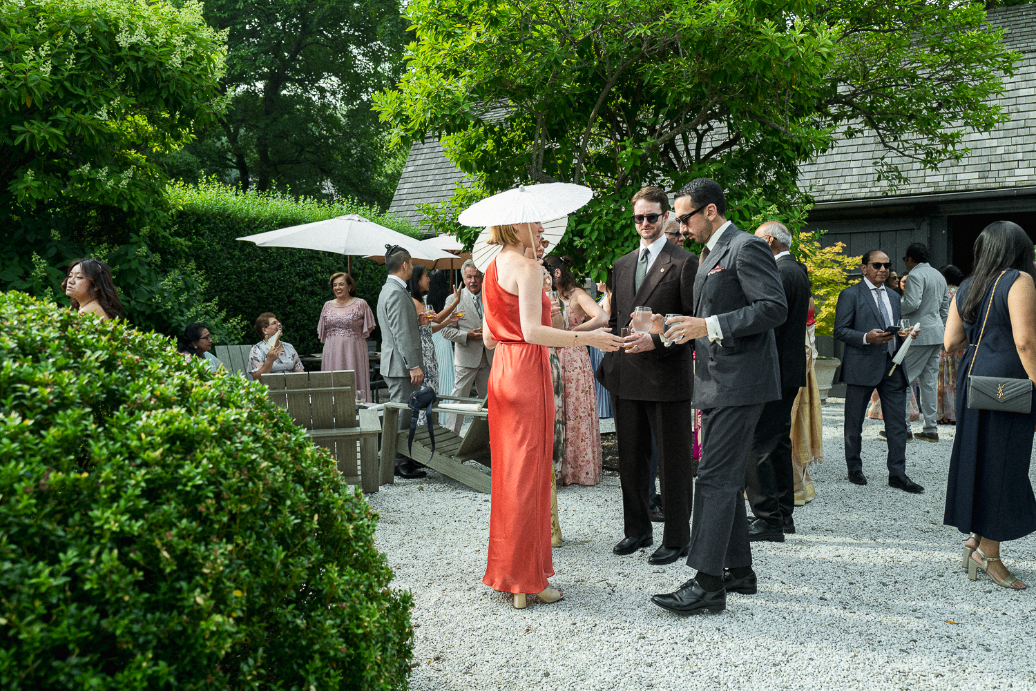 People dressed in formal attire enjoying a garden party with some holding drinks, surrounded by lush greenery and umbrellas providing shade.