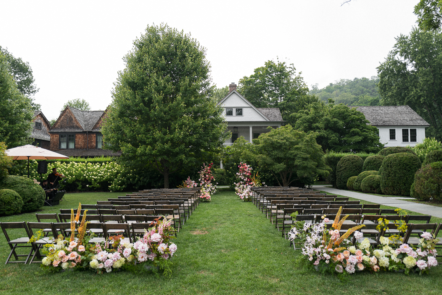 Outdoor garden setup for a wedding ceremony with rows of chairs decorated with pink and white flowers, surrounded by lush greenery and trees, with a small band playing under an umbrella on the left and houses in the background.