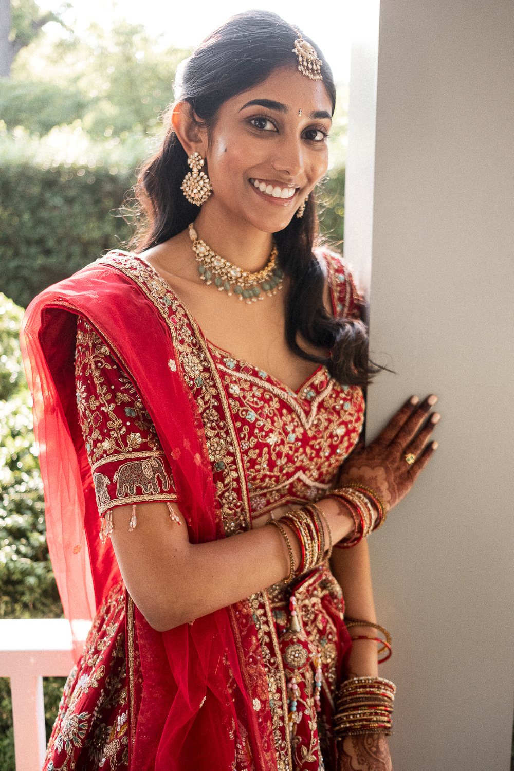 A woman dressed in traditional Indian bridal attire in red with gold embroidery, wearing jewelry including earrings, necklaces, bangles, and a headpiece, standing at an entrance with a smile.