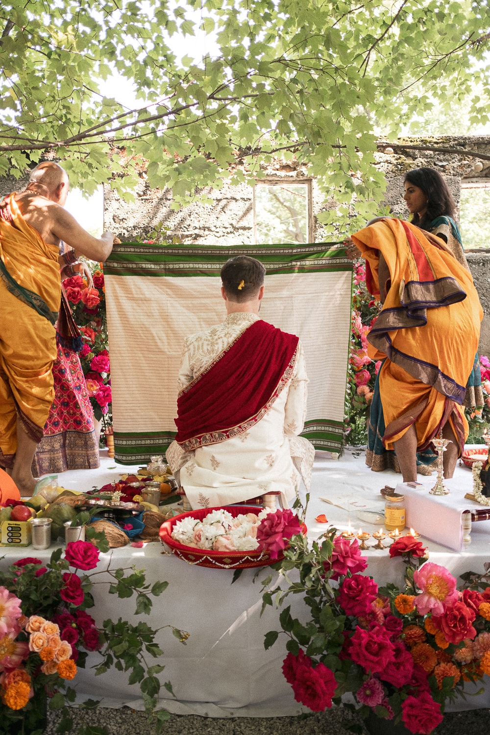 A traditional Indian wedding ceremony with a groom seated and a priest performing the rituals, surrounded by flower decorations and offerings.