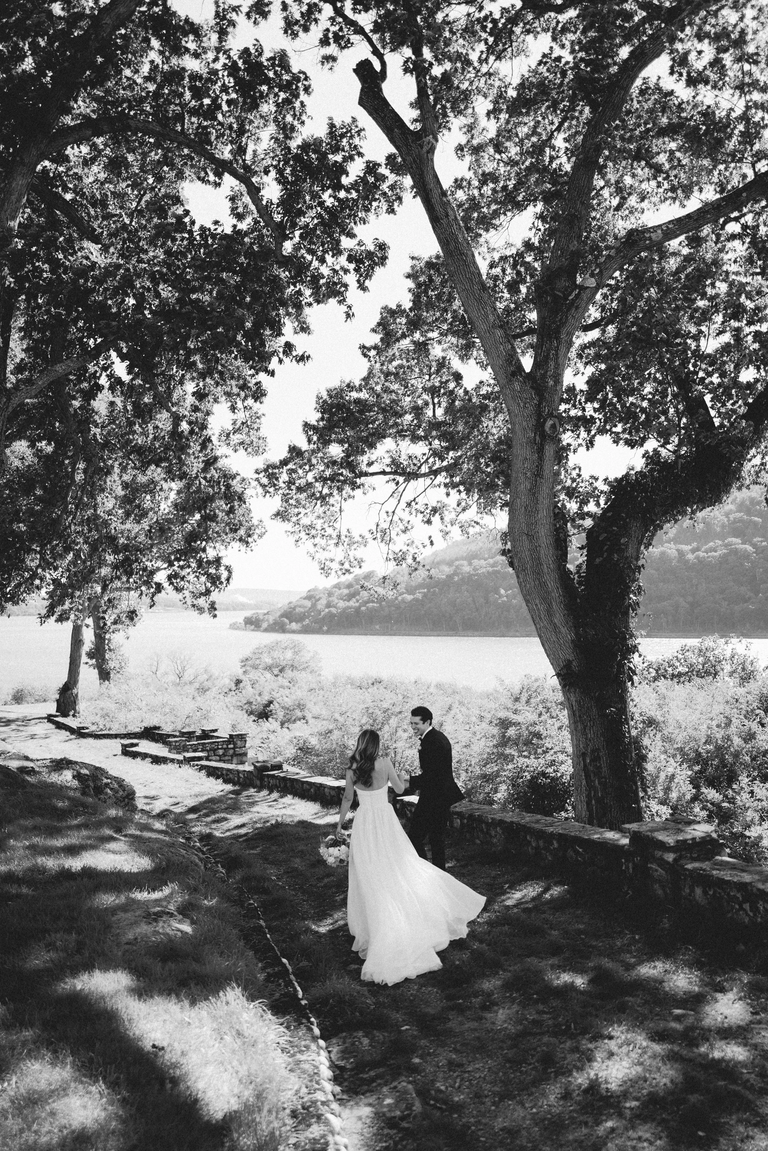 Black and white photo of a bride and groom walking near a river, under large trees, with hills and water in the background.