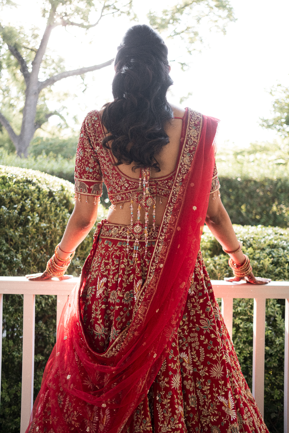A woman dressed in a red embroidered traditional Indian outfit with gold accents, standing outdoors and leaning on a white railing, with greenery and trees in the background.