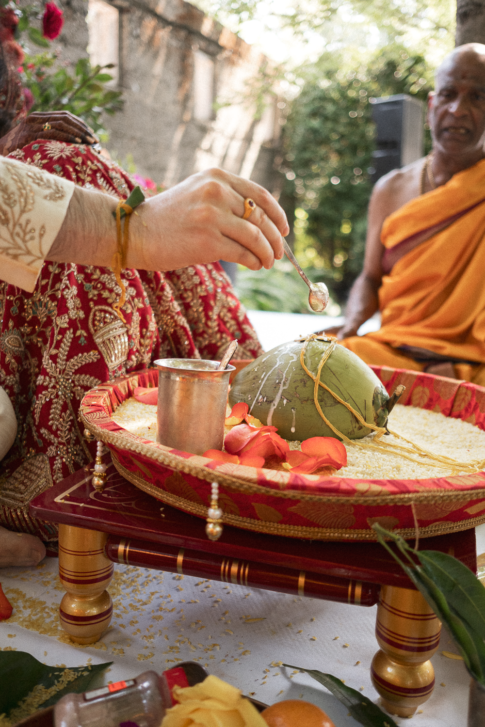 A person in traditional attire performing a ritual with a coconut, flowers, and rice on a decorated tray, with a priest or monk observing nearby during a ceremony outdoors.