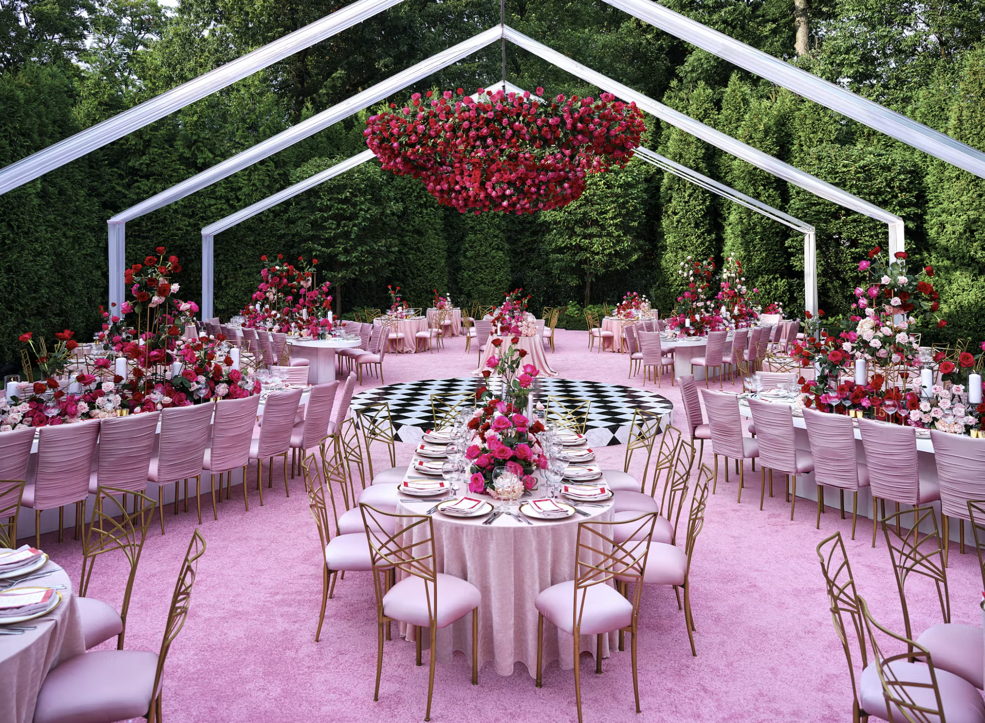 An outdoor wedding or event setup with pink carpet, round tables with pink tablecloths, elegant gold chairs, and elaborate floral centerpieces in pink and red. A large circular floral chandelier hangs from a white metal frame structure, which also su