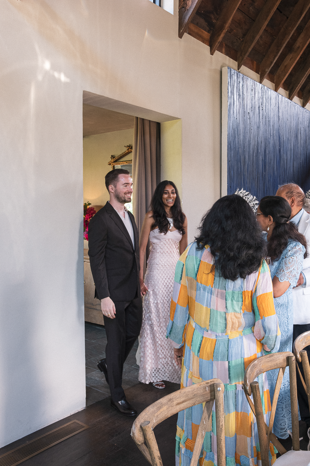A man and woman are standing in a doorway, smiling, at a social gathering. The woman is wearing a white dress, and the man is in a black suit. Other people are gathered nearby, engaged in conversation.