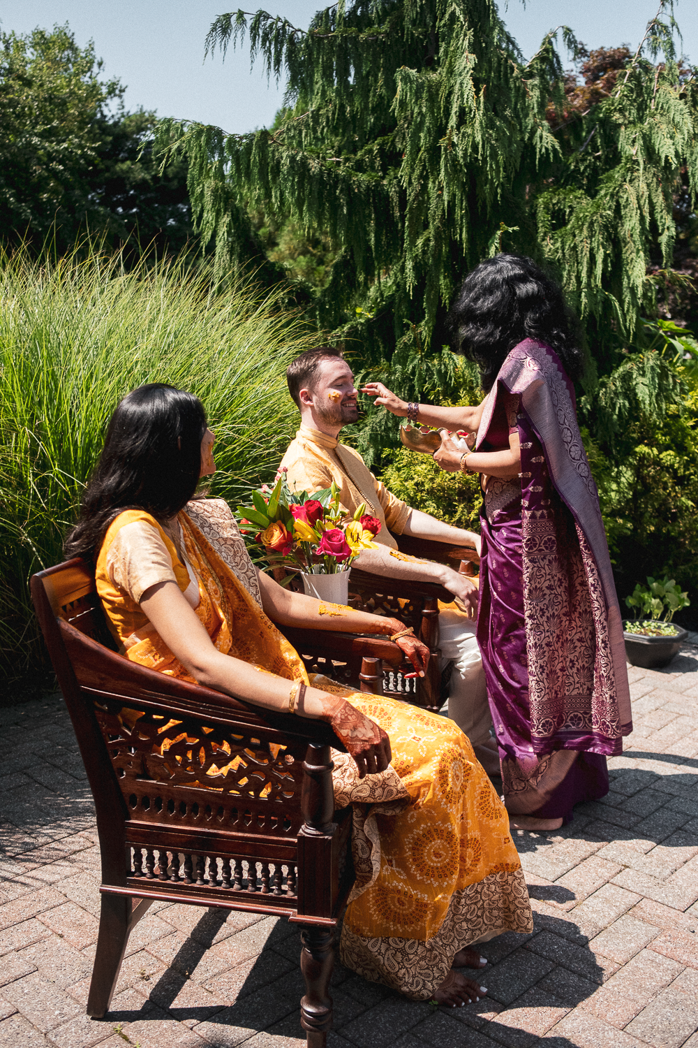 People performing a traditional Indian wedding ritual outdoors, with a woman applying a yellow powder on a man's face, both seated on wooden chairs surrounded by greenery.