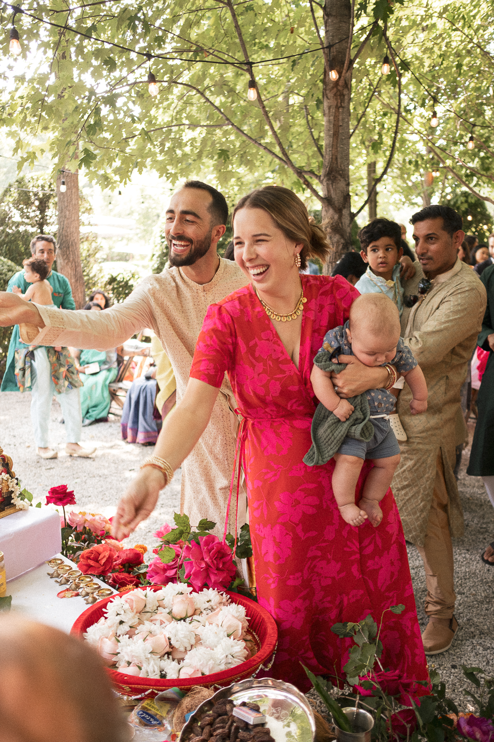 A group of people at a wedding or celebration outdoors under trees with string lights, smiling and interacting around a table with flowers and food.