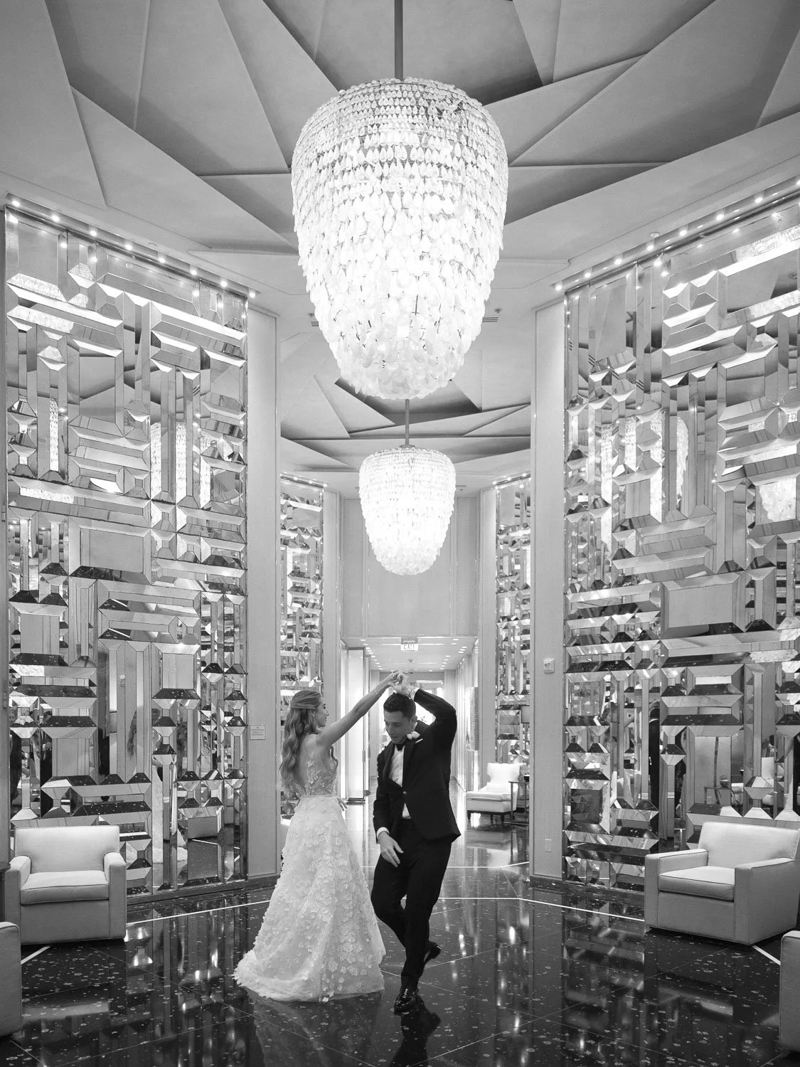 A couple dancing in an elegant, modern lobby with large chandelier lights and decorative mirrored wall panels.