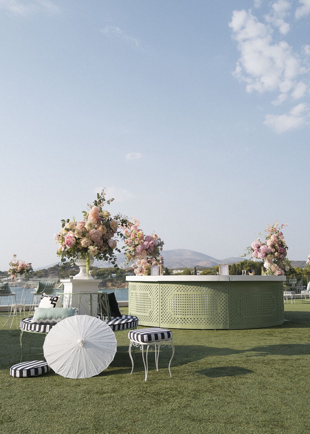 Outdoor event setup with large floral arrangements, vintage-style chairs, a green decorative bar, and a view of water and mountains in the background.