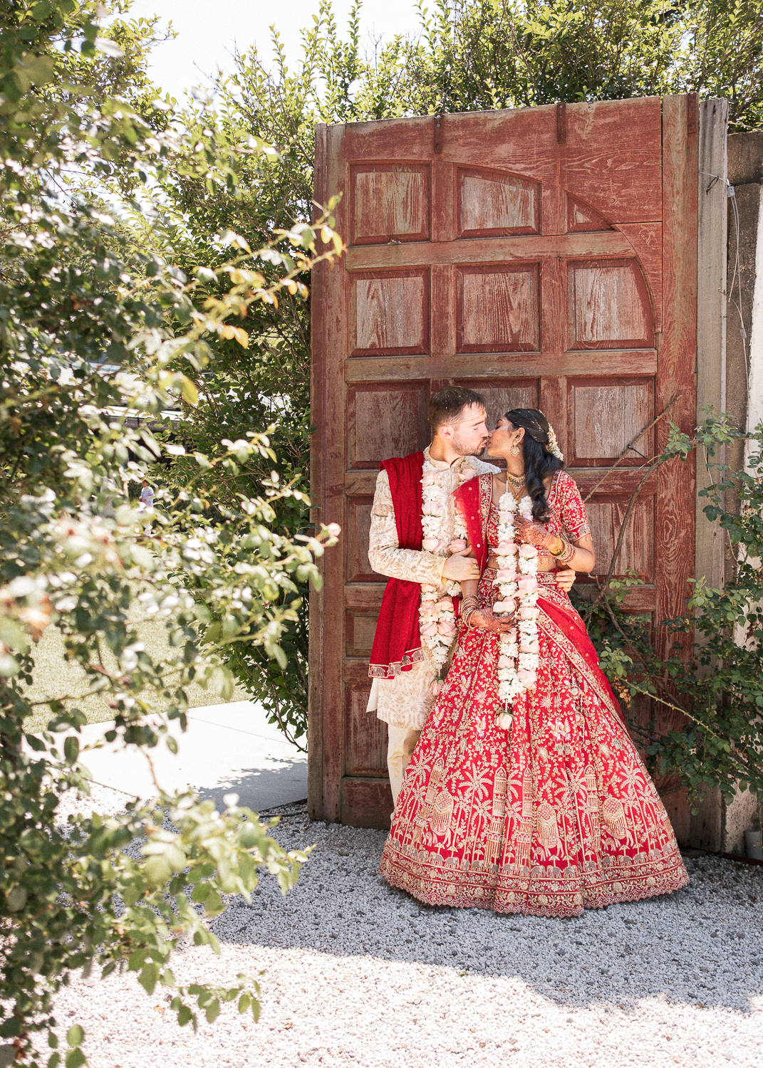 A bride and groom dressed in traditional Indian wedding attire sharing a kiss outside near a weathered wooden door.