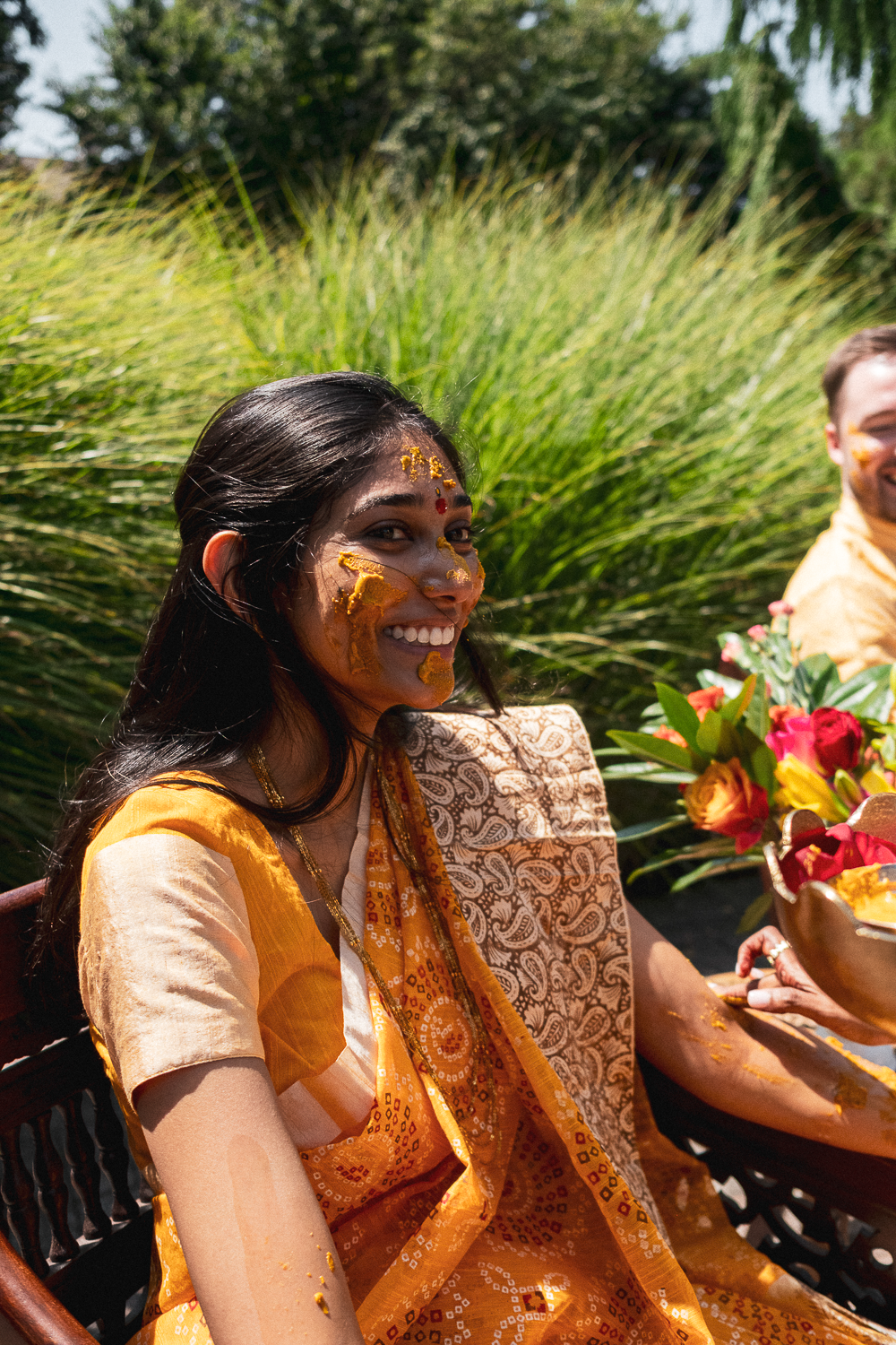 A woman smiling with turmeric paste on her face and arms during an outdoor celebration, wearing an orange traditional dress, with a man and flowers nearby.