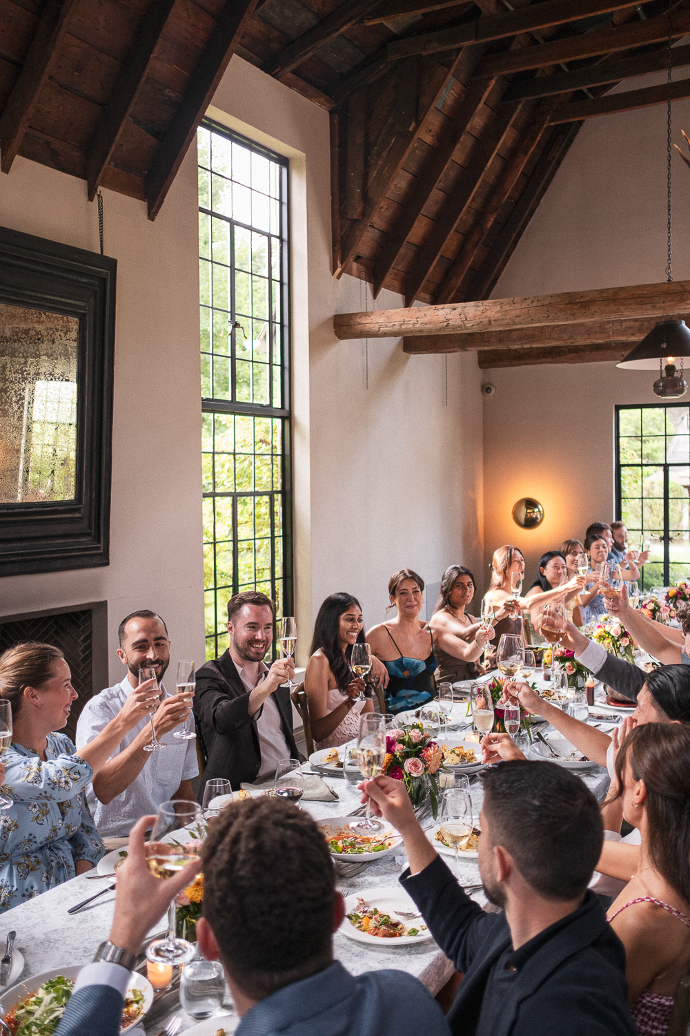 People gathered around a table raising glasses in a toast during a celebration in a rustic room with large windows and wooden beams.