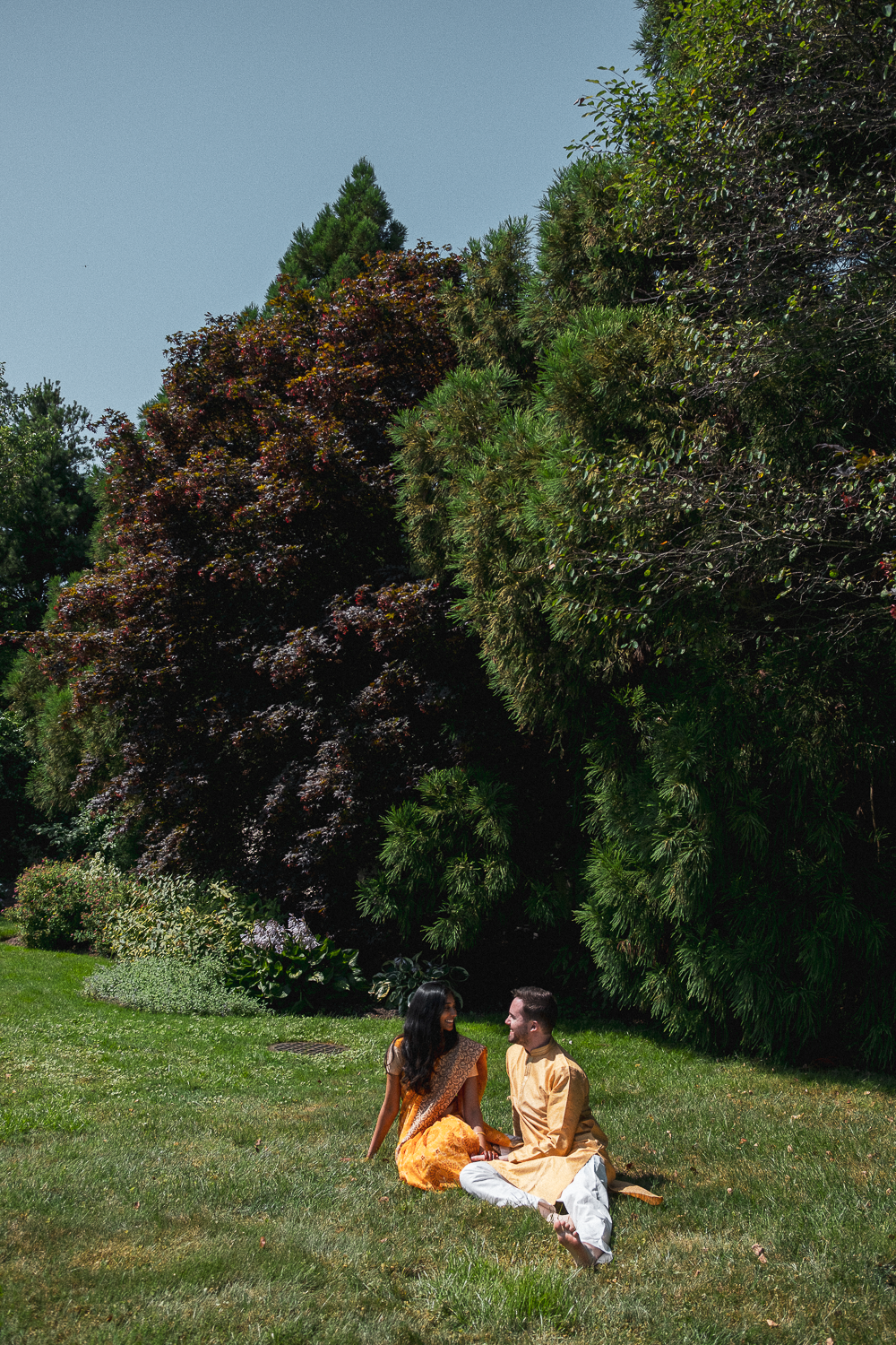 A man and woman sitting on grass in a park, dressed in traditional Indian clothing, smiling at each other, surrounded by trees and greenery.